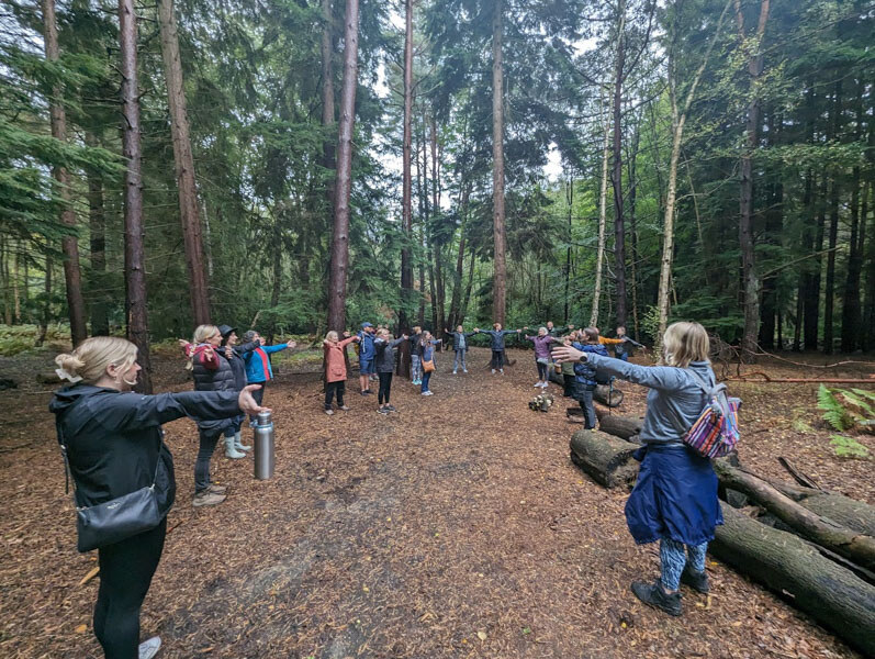 A group of people standing in a forest with their arms outstretched directly in front of them. They are all facing inwards but are not close to each other. 