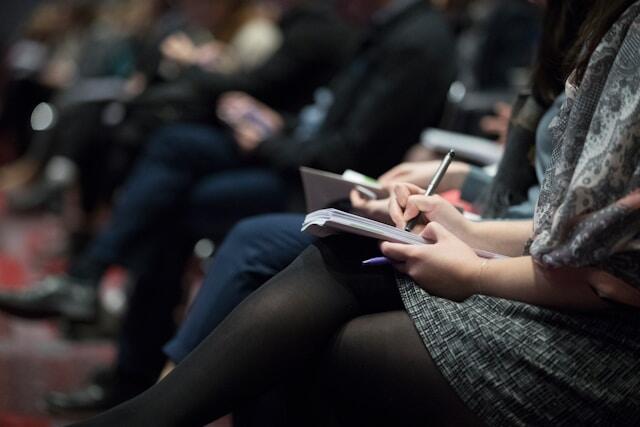 Woman sat in a conference/ theatre writing with a notepad on her lap. The image shows her side profile of her body and leg.